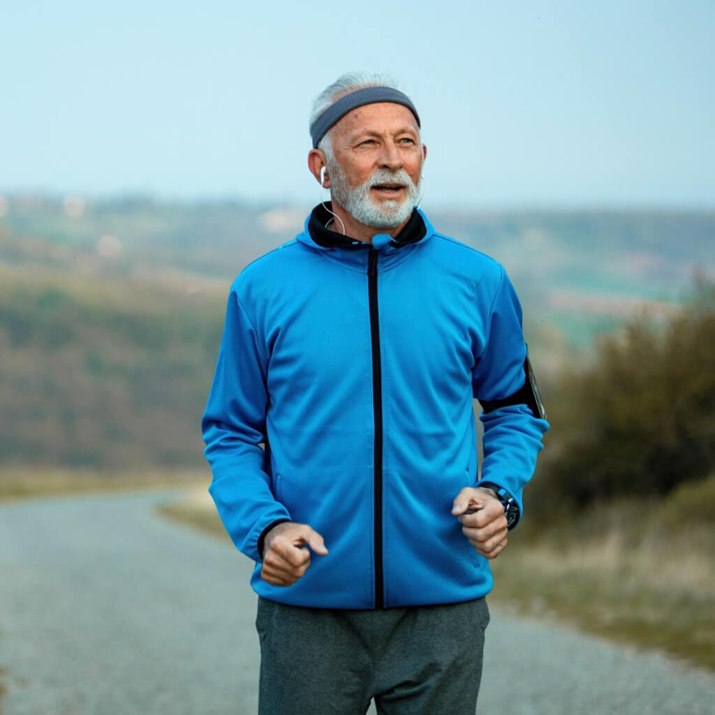 Elderly man jogging in a blue jacket on a scenic path, promoting an active lifestyle.