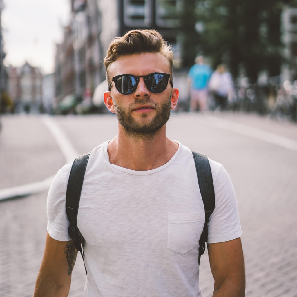 Confident man wearing sunglasses and a backpack, enjoying a sunlit day in an urban setting.