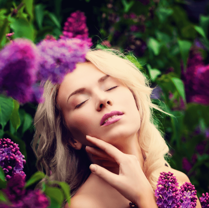 Young woman enjoying nature amidst lilac flowers, showcasing a serene expression and glowing skin.