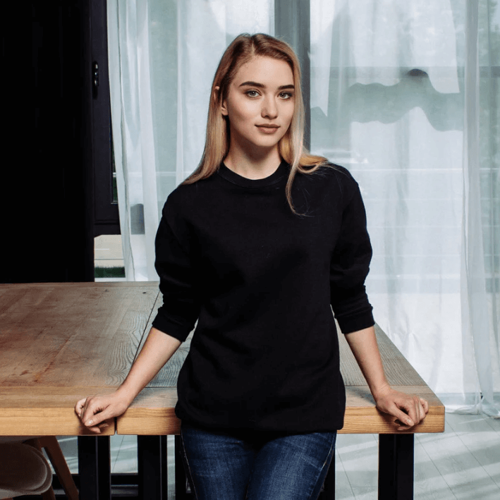 Model wearing a black sweater, posing beside a wooden table in a bright room.