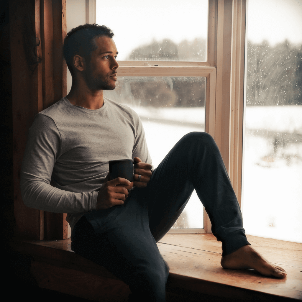 A man sitting on a windowsill, sipping tea from a mug, gazing out at a snowy landscape.
