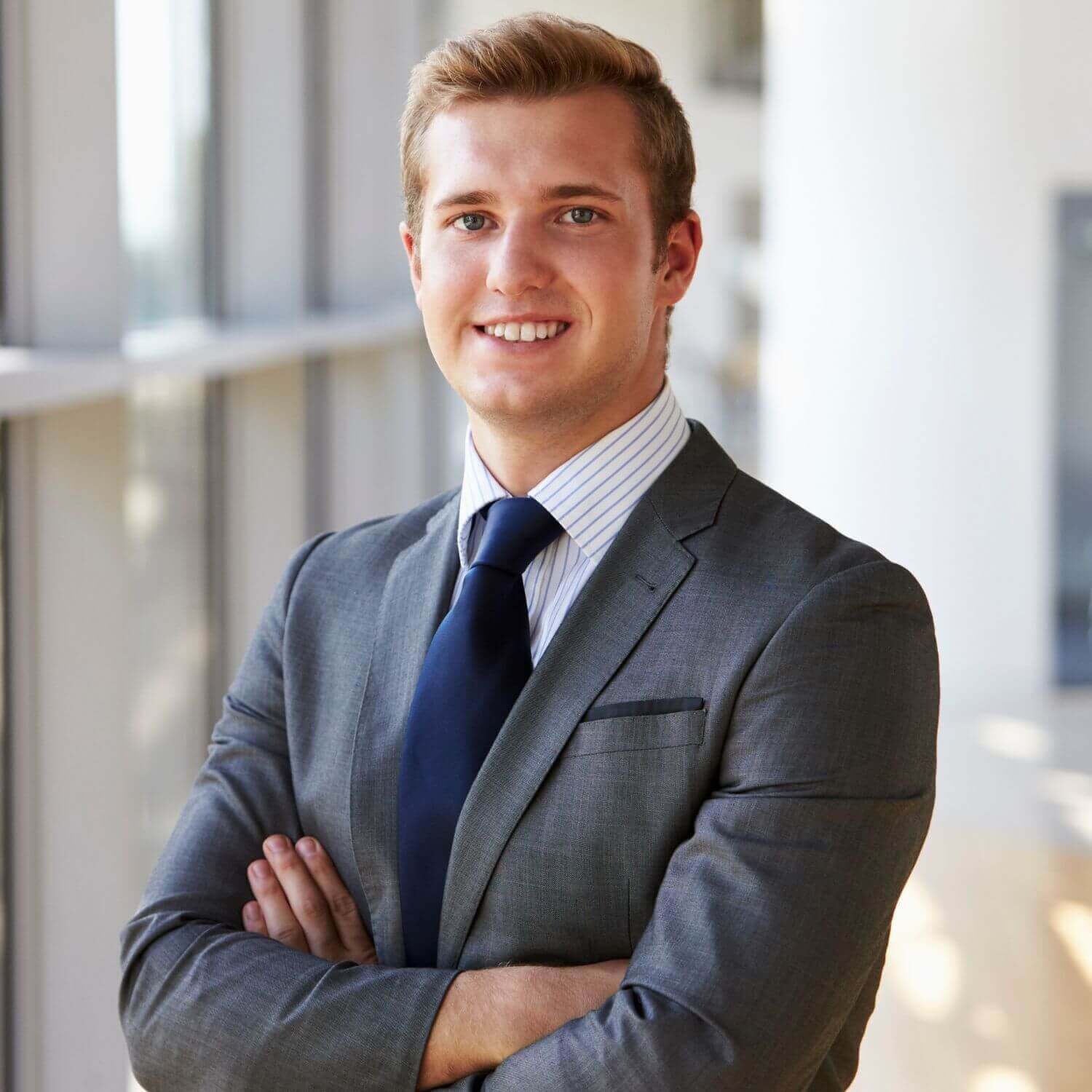 Confident young man in a business suit, smiling and posing with arms crossed in a modern office setting.