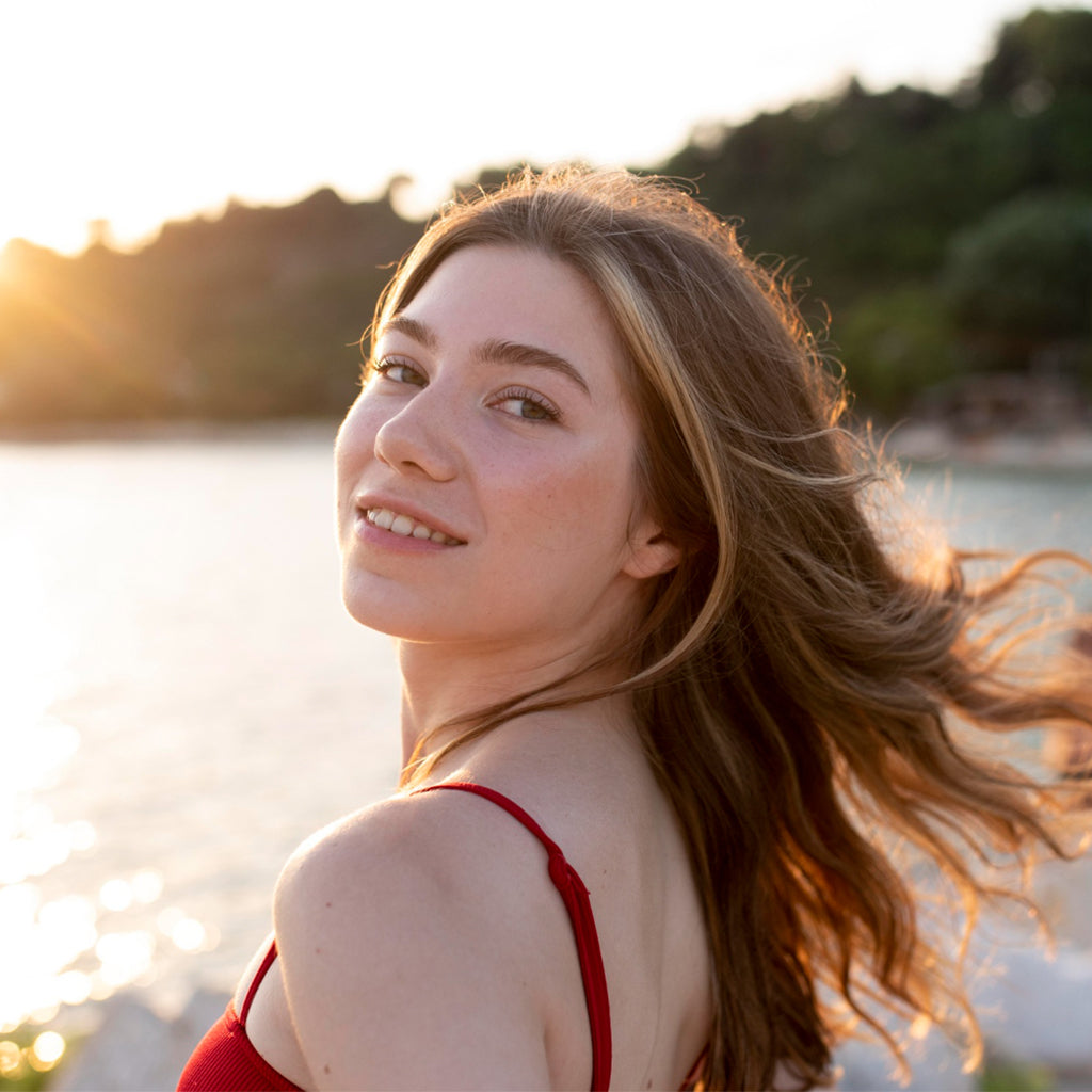 A woman with long hair, smiling at the camera, near a sunlit beach, showcasing a fresh and healthy appearance.