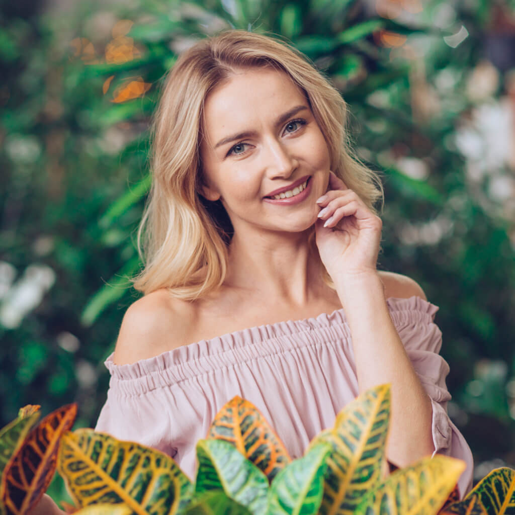 Smiling woman with blonde hair showcasing Ultra-Fine Honey Lifting Cream surrounded by lush greenery.